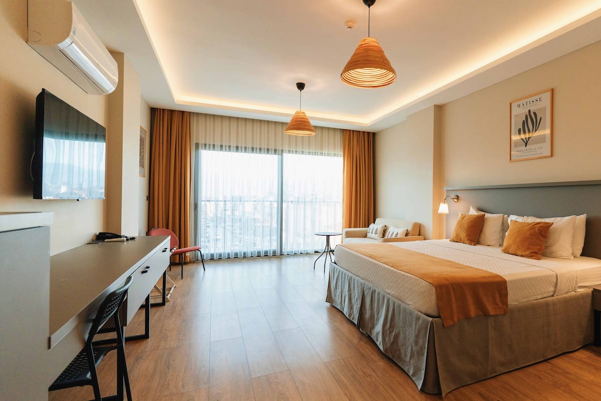 A hotel room with a bed, bedside tables, a desk, and a chair. There is a large window with curtains, a ceiling light, and a wall-mounted TV.