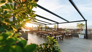 Una terraza en la azotea con mesas y sillas de madera, plantas en macetas y vistas de la ciudad.