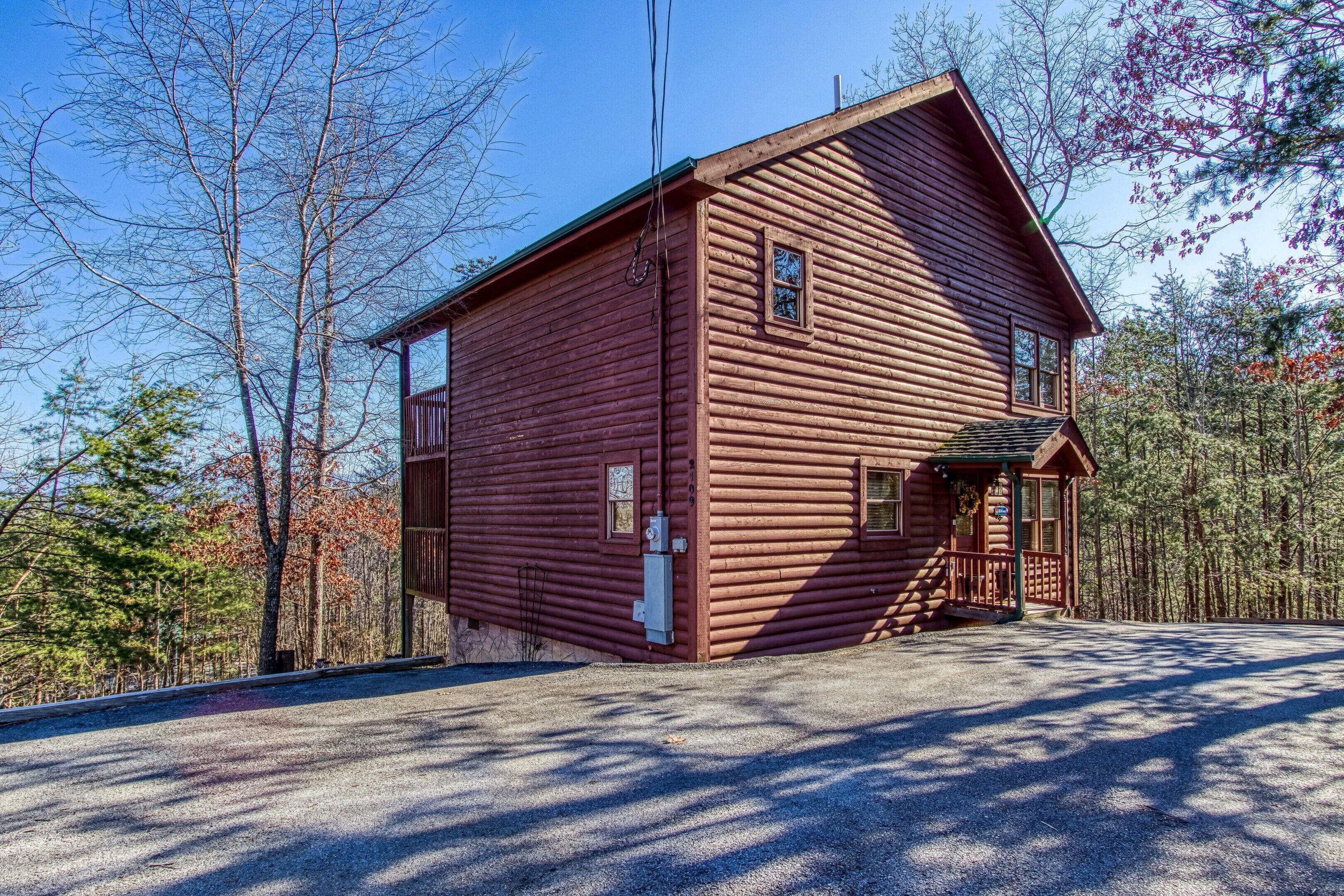 A two-story log cabin with a front porch, a small attached garage, and a gravel driveway.