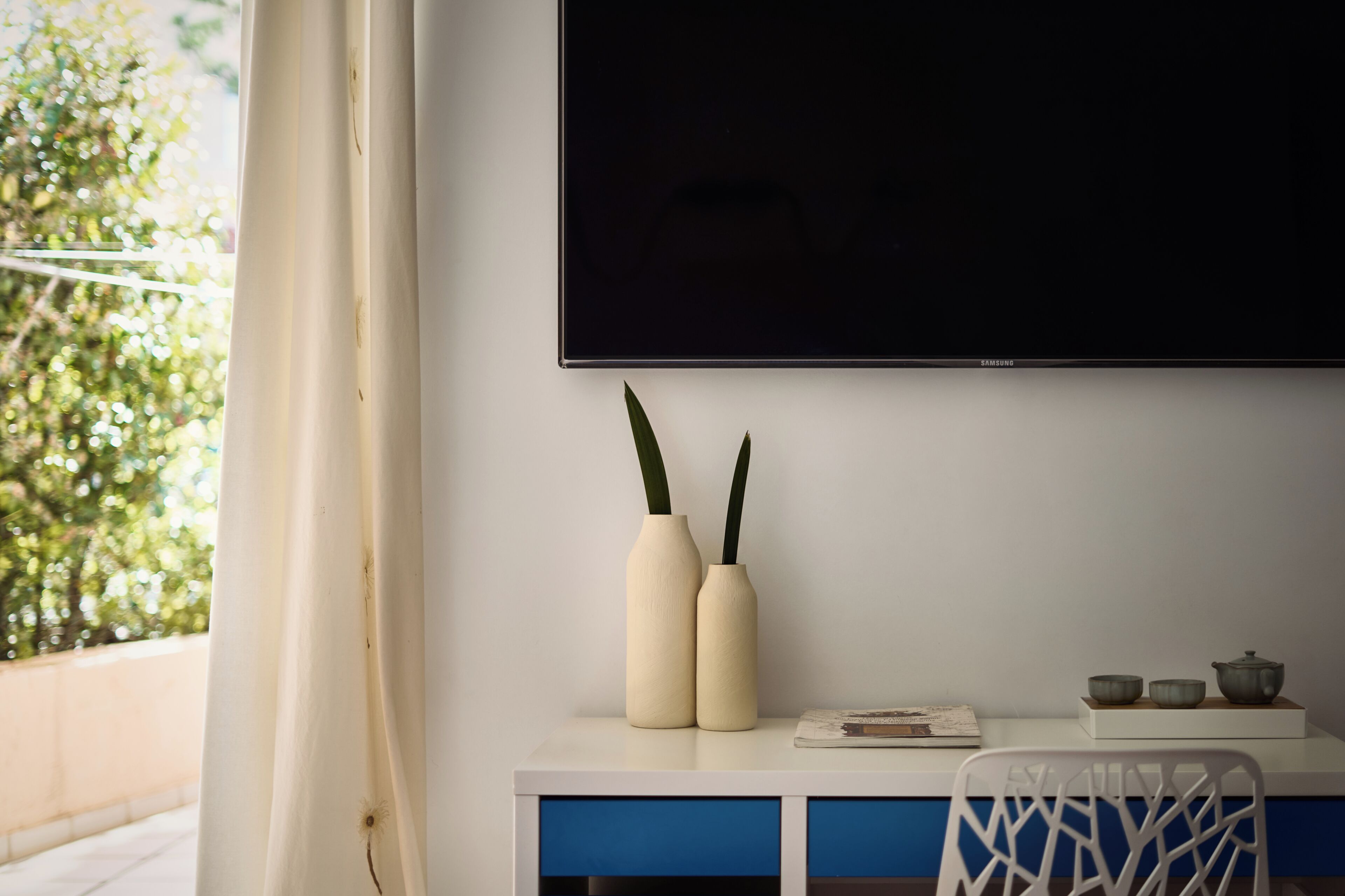A room with a flat-screen TV mounted on the wall, a white desk with blue drawers, two vases, and a chair.