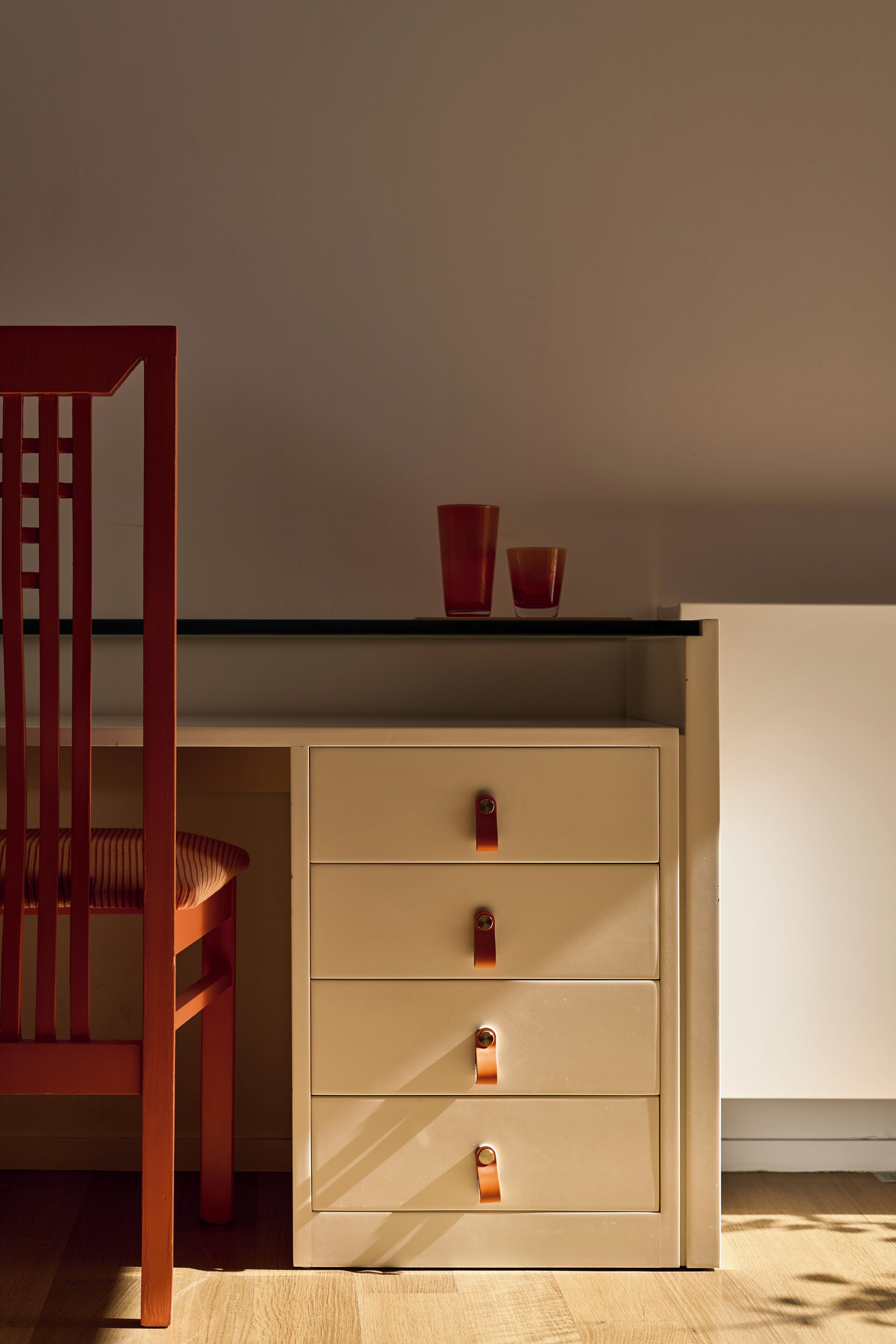 A room with a wooden chair, a beige cabinet with drawers, and a shelf with two red cups.