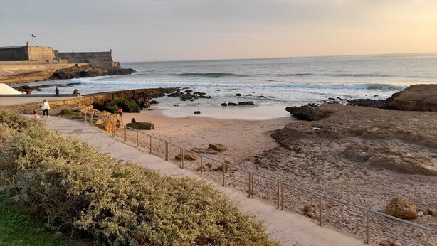 A coastal walkway leading to a sandy beach with rocky outcrops, a flag-flagged building, and a clear sky. - Carcavelos Surf Beach House (Carcavelos)