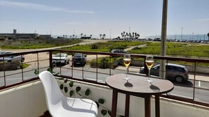A balcony with a white chair and a small round table, overlooking a parking area and a beach with palm trees. - Carcavelos Surf Beach House (Carcavelos)