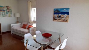 A modern living room with a glass dining table, white chairs, a sofa with orange cushions, and a beach-themed painting on the wall. - Carcavelos Surf Beach House (Carcavelos)