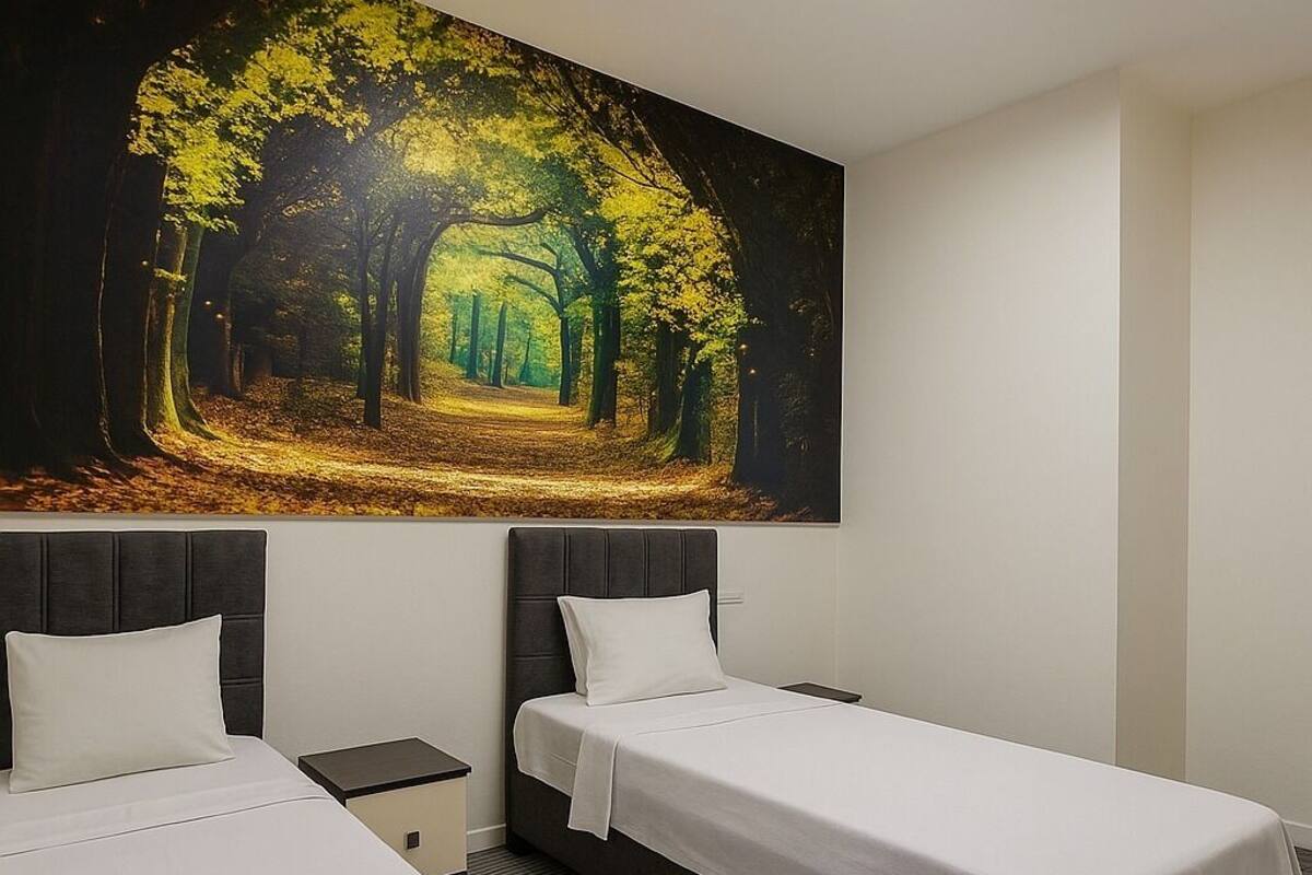 A hotel room with two beds, a large forest-themed painting on the wall, and a skylight above.