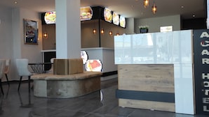A reception area with a reception desk, seating area with chairs, and a wall-mounted TV displaying food images. - Acar Airport Hotel Plus (Arnavutköy)