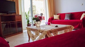 Un salon avec un canapé rouge, un téléviseur posé sur un meuble en bois, une table recouverte d’une nappe en dentelle et des plantes en pot.