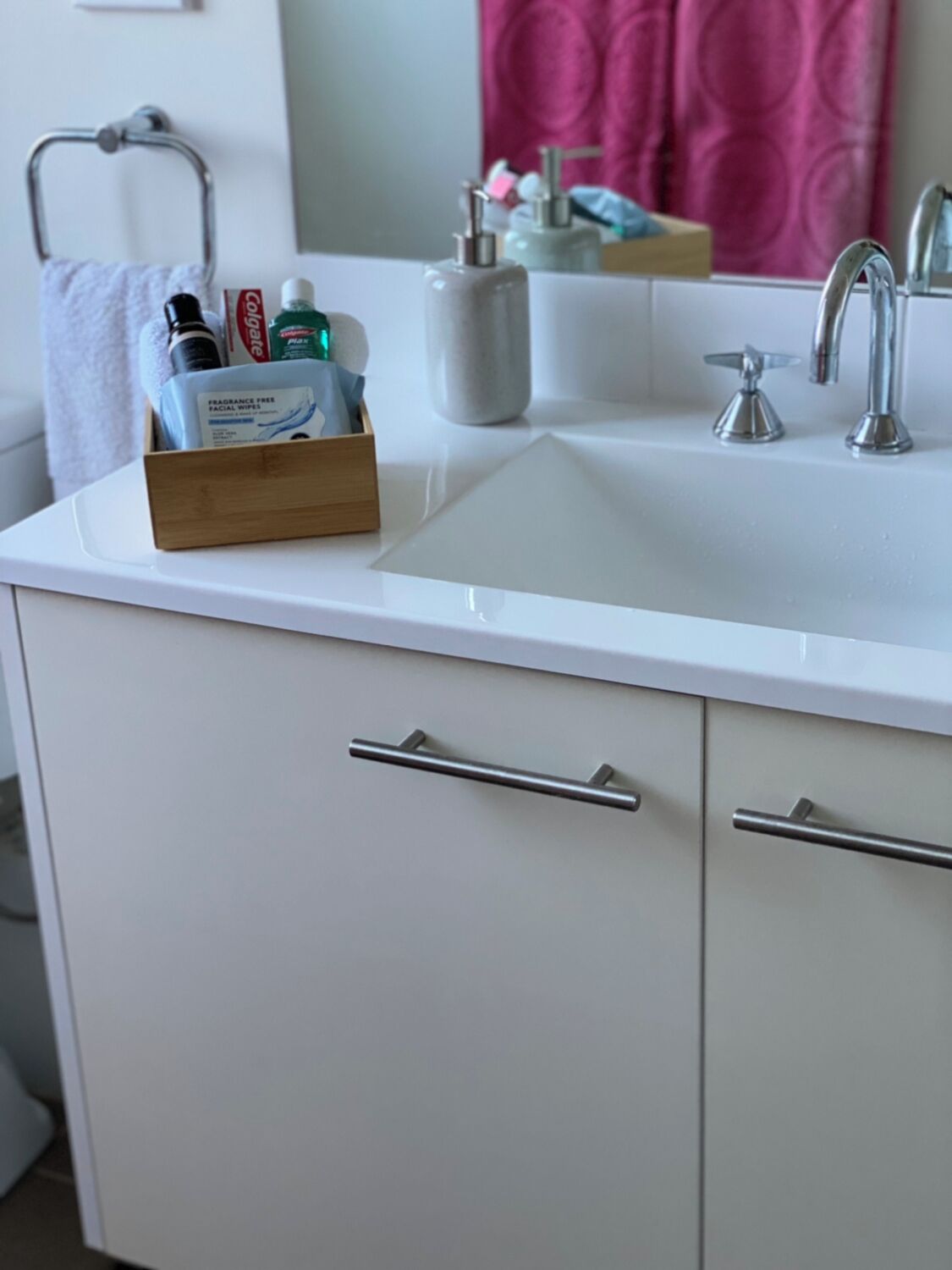Une salle de bain avec un lavabo blanc, un miroir et un porte-serviettes. On trouve une boîte en bois contenant des articles de toilette sur le lavabo.