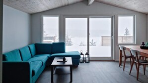 Una sala moderna con un sofá color esmeralda, una mesa de comedor de madera con sillas y vistas a un paisaje nevado a través de grandes puertas correderas de vidrio.