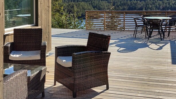 Una terraza de madera con mobiliario de mimbre, incluyendo sillas y una mesa, con vistas a un área boscosa.