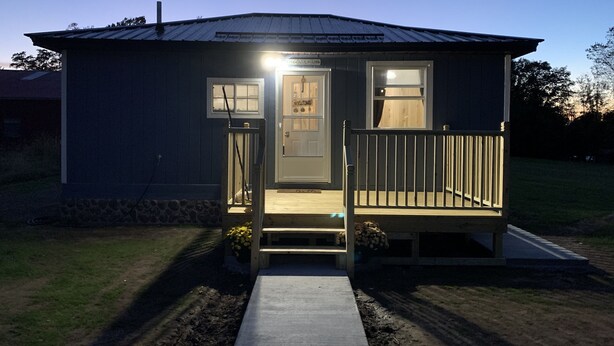 A small, single-story building with a porch, a welcome sign, and a door. - Two bedroom Cottage (Cornell)