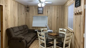 A cozy room with a brown sofa, a dining table with white chairs, a ceiling fan, and wooden walls. - Two bedroom Cottage (Cornell)
