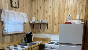 A kitchen with wooden panel walls, a white refrigerator, a stove, a sink with a checkered mat, and a clock on the wall. - Two bedroom Cottage (Cornell)