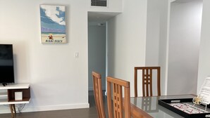 A dining area with a glass-top table, wooden chairs, a flat-screen TV, and a Miami Beach poster on the wall. - Blocks Aways From Brickell Village, 2 beds 2 Baths FREE Parking NO Cleaning FEES (Miami)