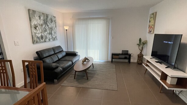 A modern living room with a black leather sofa, a glass coffee table, a dining table with chairs, a flat-screen TV, and a large window with blinds. - Blocks Aways From Brickell Village, 2 beds 2 Baths FREE Parking NO Cleaning FEES (Miami)