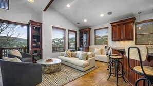 A spacious living area with a sofa, coffee table, and bar stools. There are large windows offering a view of the landscape. - 502 A Gold Coast! 3 Bedroom Townhouse by RedAwning (Park City)