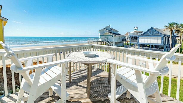 Una terraza de madera con sillas blancas y una mesa, con vistas a una playa y casas al fondo.