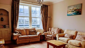 A cozy living room with a sofa, armchair, ottoman, and a painting on the wall. - Huge 4 Bed Apt - Perfect Base to Explore The Borders (Jedburgh)