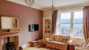 A cozy living room with a fireplace, a sofa, a wooden coffee table, and a television mounted on the wall. - Huge 4 Bed Apt - Perfect Base to Explore The Borders (Jedburgh)