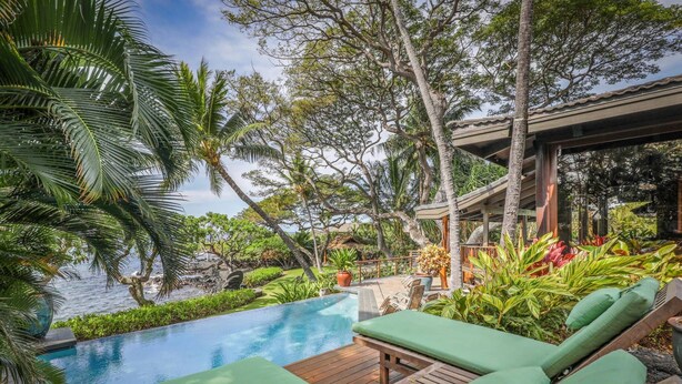 Una piscina infinita con terraza de madera y sillones reclinables verdes, rodeada de plantas tropicales y palmeras.