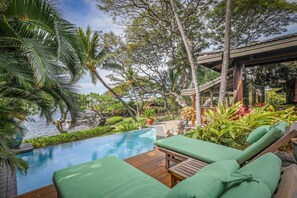 An infinity pool with wooden deck and green lounge chairs, surrounded by tropical plants and palm trees.