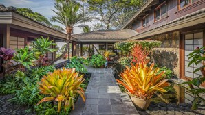 Un edificio de estilo tradicional con un paseo cubierto, rodeado de exuberante vegetación tropical y una pequeña fuente de agua.