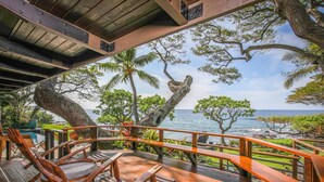 Una terraza de madera con sillas y una mesa, con vista a una playa con palmeras y el océano.