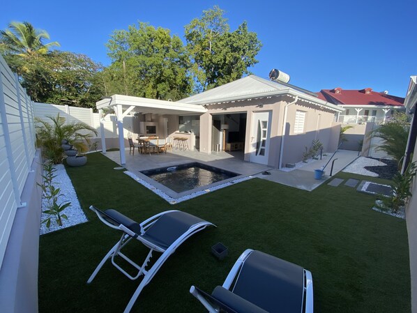 Un patio trasero con piscina, dos sillas de descanso, una mesa de patio y una zona de estar techada.