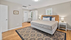 Une chambre avec un grand lit, une table de chevet avec une lampe, un tapis à motifs et une porte donnant sur une autre pièce.