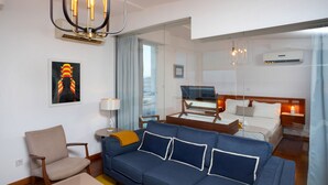 A modern hotel room with a blue sofa, a wooden side table, a lamp, and a bed with a wooden headboard. - 1-80 Collection Penthouse by TrulyCyprus (Limassol)