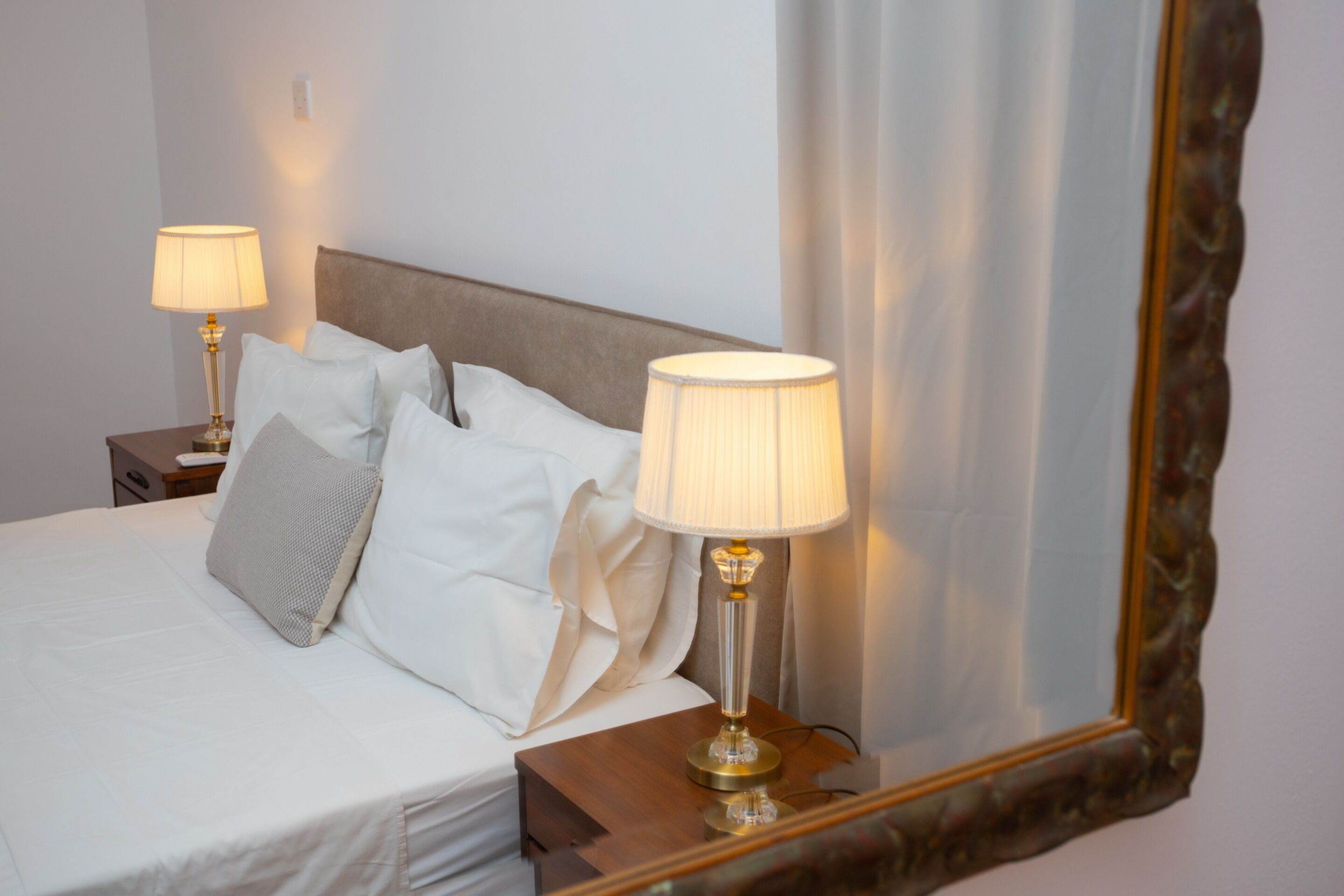A neatly made bed with white linens and pillows, a bedside table with a lamp, and a large mirror with a decorative frame.