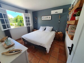 A bedroom with a bed, bedside tables, a window with curtains, a wall-mounted air conditioner, and a bookshelf. - Villa with sea view and close to rivers, 2mn from Valombreuse floral park (PETIT-BOURG)