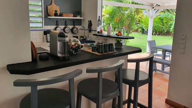 A modern kitchen with a bar area, featuring a coffee maker, a kettle, and a fruit bowl. There are two bar stools and a view of a garden through the open door. - In the heart of the island, with a view of the sea, near the rivers, 2 minutes from the Vallombreuse Garden (PETIT-BOURG)