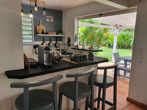 Eine moderne Küche mit Barbereich, ausgestattet mit einer Kaffeemaschine, einem Wasserkocher und einer Obstschale. Zwei Barhocker und Blick auf einen Garten durch die offene Tür.