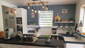 A modern kitchen with a central island, bar stools, a window with blinds, hanging pots, and a clock on the wall. - In the heart of the island, with a view of the sea, near the rivers, 2 minutes from the Vallombreuse Garden (PETIT-BOURG)