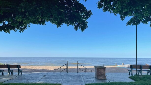 A paved walkway leading to a beach with benches and a trash bin.