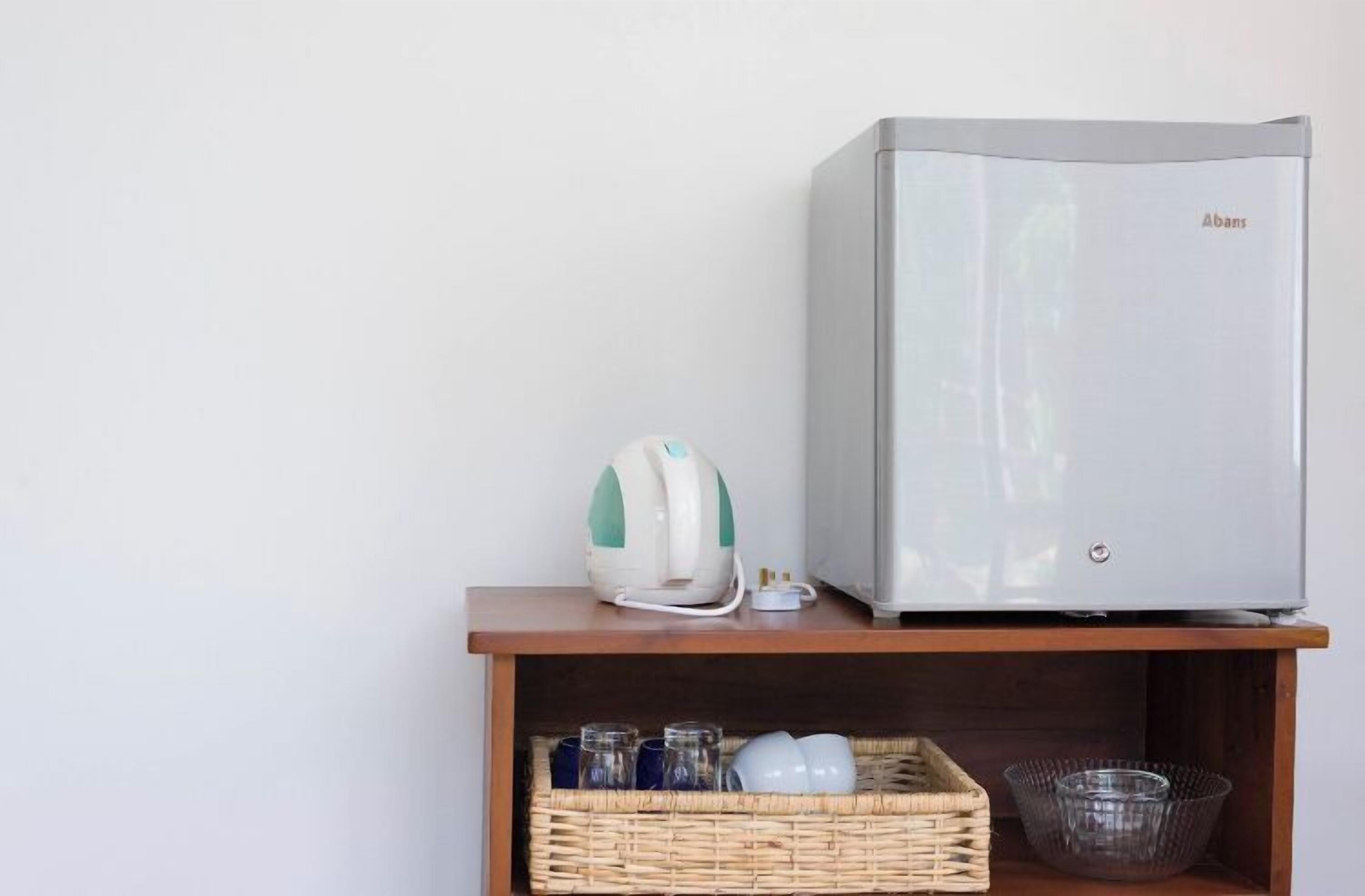 A compact kitchenette with a modern refrigerator, a kettle, and a wicker basket containing glasses and dishes.