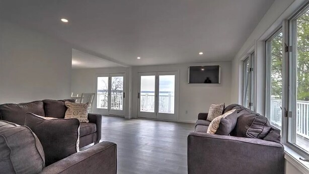 Uma sala de estar espaçosa com sofás cinzas, uma televisão de tela plana fixada na parede e grandes janelas com vista para o exterior.