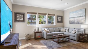 Ein Wohnzimmer mit grauem Sofa, einem Holztisch und einem Fernseher auf einem Holzgestell. Es gibt drei Fenster mit Jalousien, einen Deckenventilator und ein Bild an der Wand.