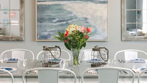 Ett matbord med vita stolar, en vas med blommor och en tavla med ett havsmotiv på väggen.