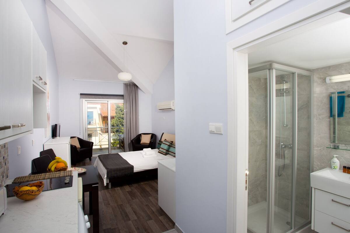 Ein kompaktes Hotelzimmer mit Bett, kleinem Schreibtisch, Stuhl, Esstisch mit Obstschale, Duschbereich sowie Bad mit Spiegel und Waschbecken.