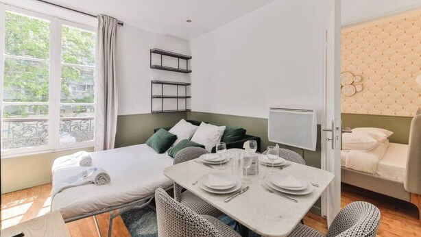 A small, well-lit room with a bed, a dining table set for two, and a balcony with a view of greenery.