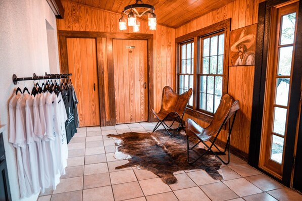 A room with wooden walls, a fur rug, two leather chairs, a coat rack with hanging clothes, and a painting on the wall. - The Vaquero Motel (Bandera)