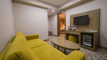 A hotel room with a yellow sofa, a small round table, a television mounted on the wall, and a minibar.