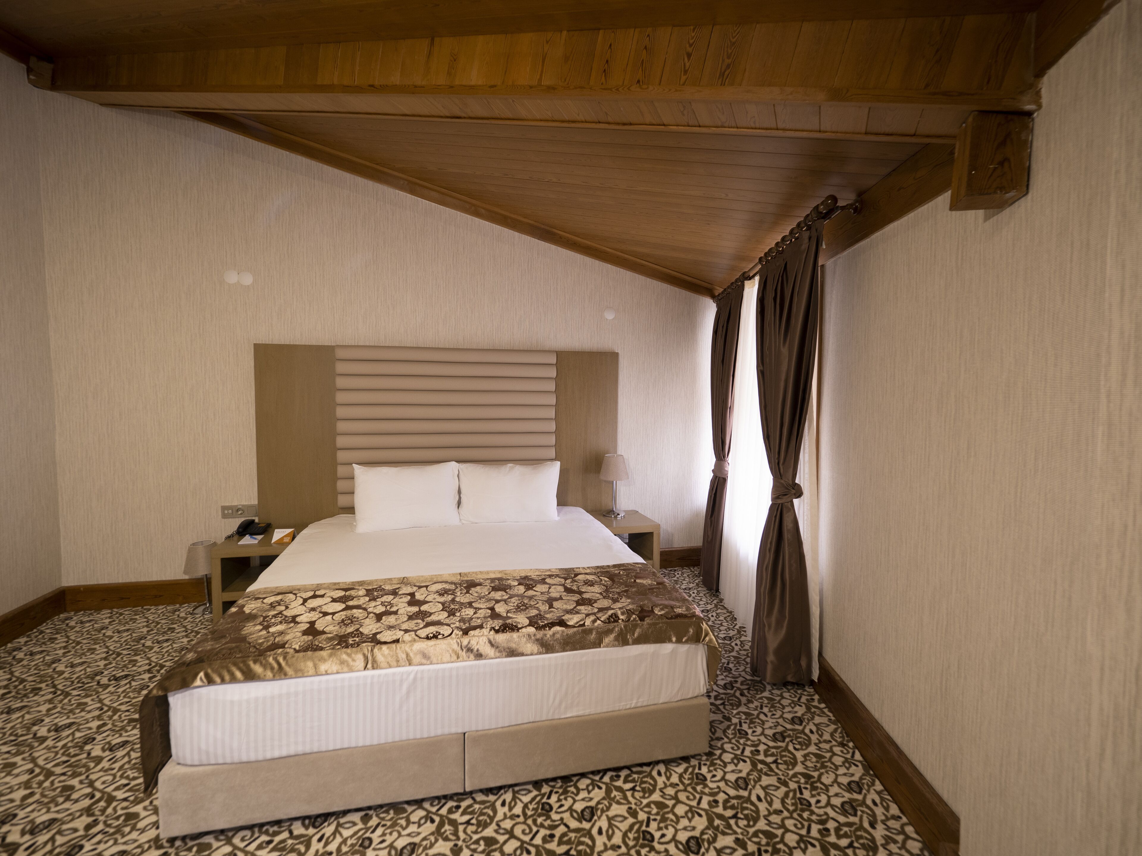 Een slaapkamer met een houten plafond, een bed met een beddeken met dessin, een nachtkastje met een lamp en een tapijt met patroon.