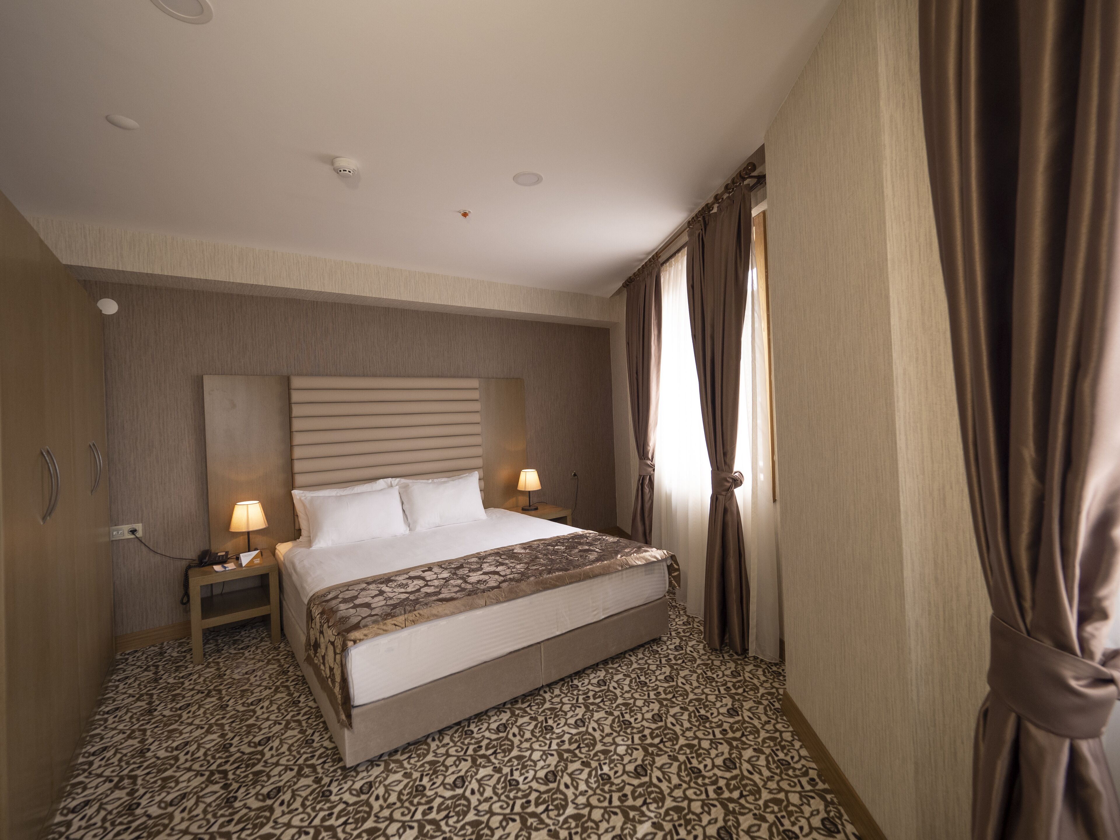 Habitación de hotel con una cama grande, dos lámparas de noche, un cabecero, una mesita de noche y una alfombra estampada.