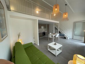 A modern living room with a green sofa, a white coffee table, and a dining area with a table and chairs. There is a staircase leading to an upper level.