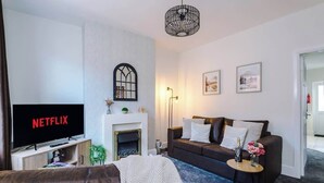 A living room with a television displaying 'NETFLIX', a fireplace, a sofa, and a coffee table with flowers. - Fresh house, amazing location, great for couples! (Chester)