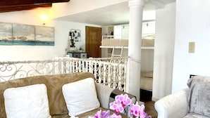 A cozy living room with a brown sofa, white cushions, and a decorative orchid plant. A white railing separates the living area from a bedroom with a bunk bed. - Duplex house with garden in the hills above Cagnes  (Cagnes-sur-Mer)