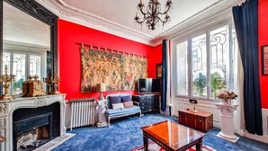 A room with a red wall, a large tapestry, a fireplace, a blue sofa, a TV, and a table. - #Arc DeTriomphe #Trocadero #Style Haussmanien#AC (Paris)